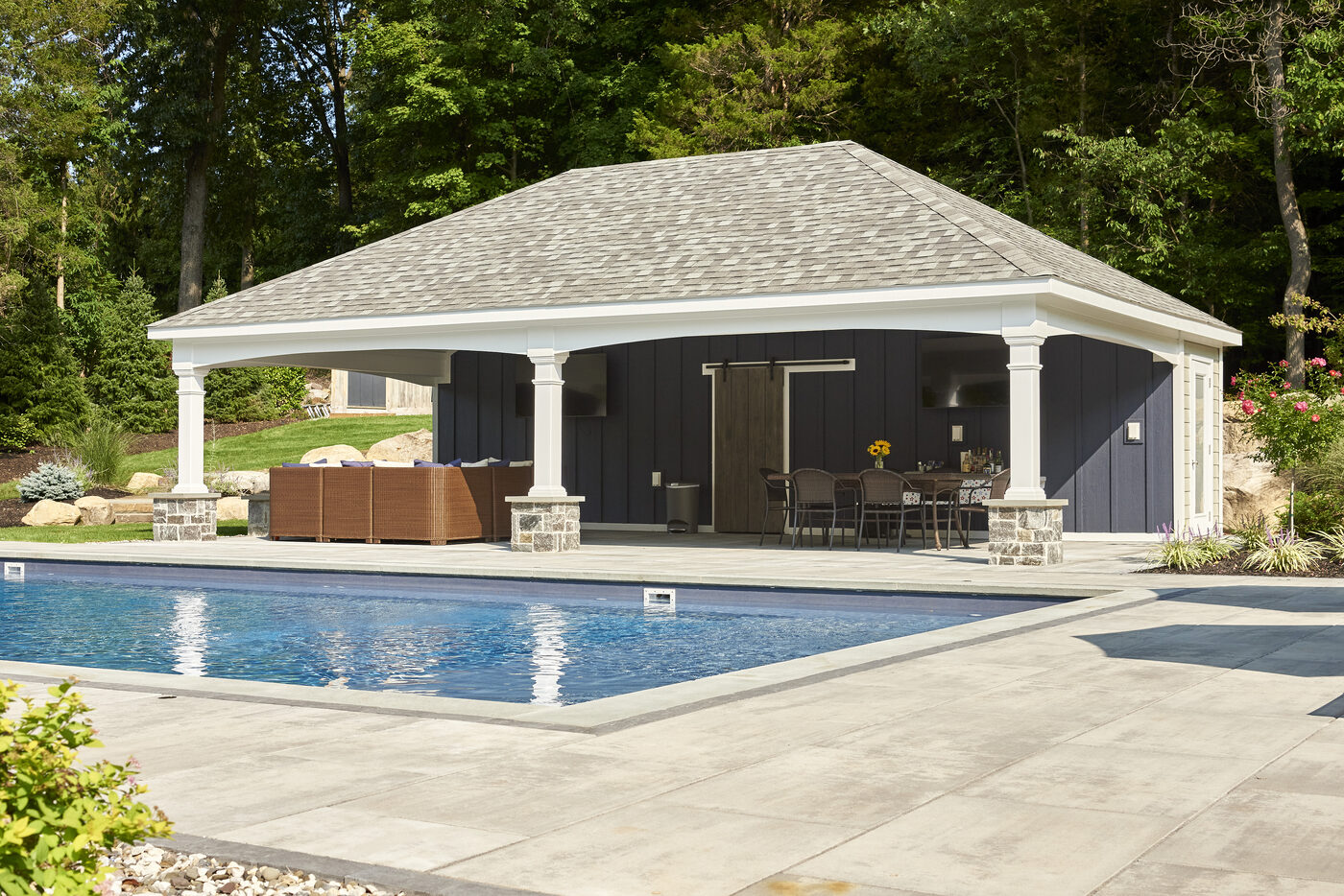 Homestead Avalon Hip Roof pool house with cape cod vinyl dutch lap siding, matte black standing seam metal roof, illuminated at dusk beside a pool